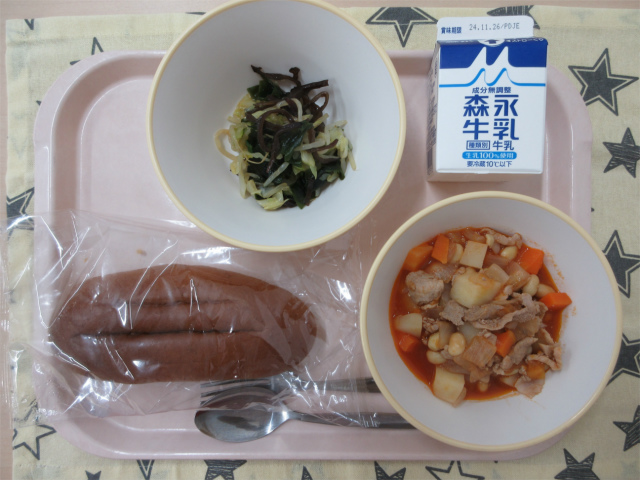 ピンクのトレーに給食が右上から反時計回りで牛乳、海藻サラダ、黒パン、ポークビーンズと並べられている写真