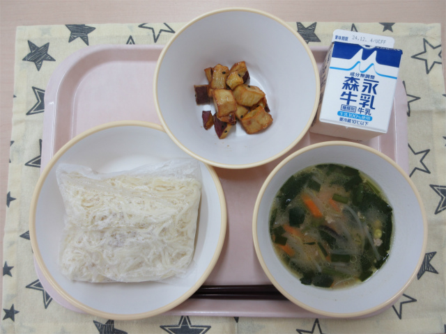 ピンクのトレーに給食が右上から反時計回りで牛乳、大学芋、袋入りの麺、味噌ラーメンのスープと並べられている写真