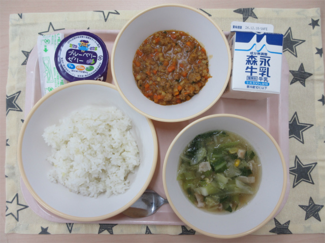 ピンクのトレーに給食が右上から反時計回りで牛乳、根菜入りキーマカレー 、ブルーベリーゼリー、ごはん、キャベツのスープと並べられている写真