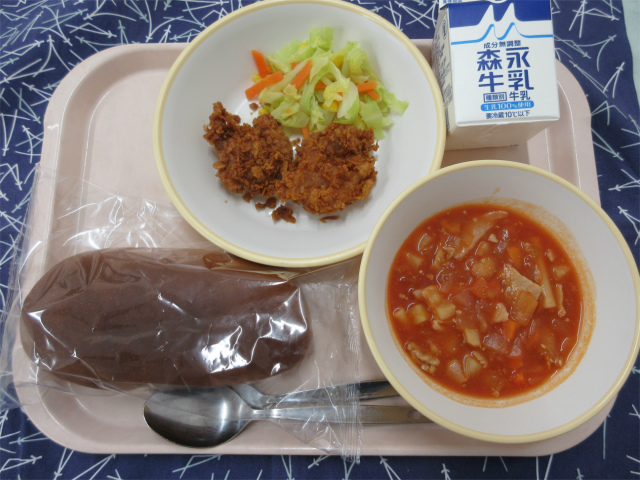 ピンクのトレーに給食が右上から反時計回りで牛乳、クリスピーチキンとキャベツソテー、黒パン、ミネストローネと並べられている写真