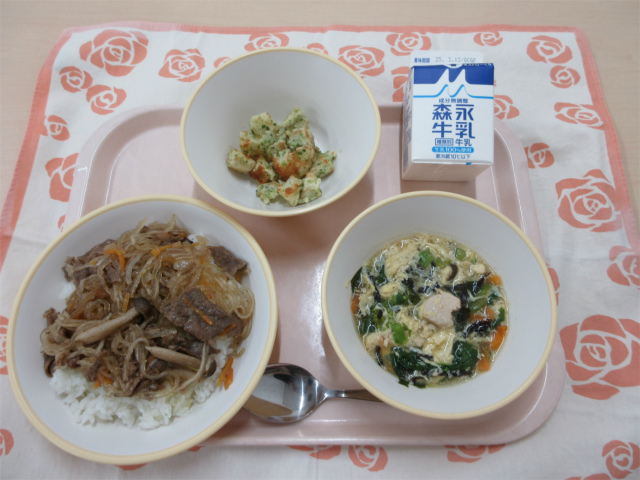 ピンクのトレーに給食が右上から反時計回りで牛乳、青のりポテト、チャプチェ丼、卵とわかめスープと並べられている写真