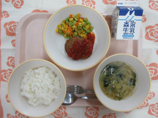 ピンクのトレーに給食が右上から反時計回りで牛乳、ハンバーグトマトソースとカラフルソテー、ごはん、キャベツのスープと並べられている写真