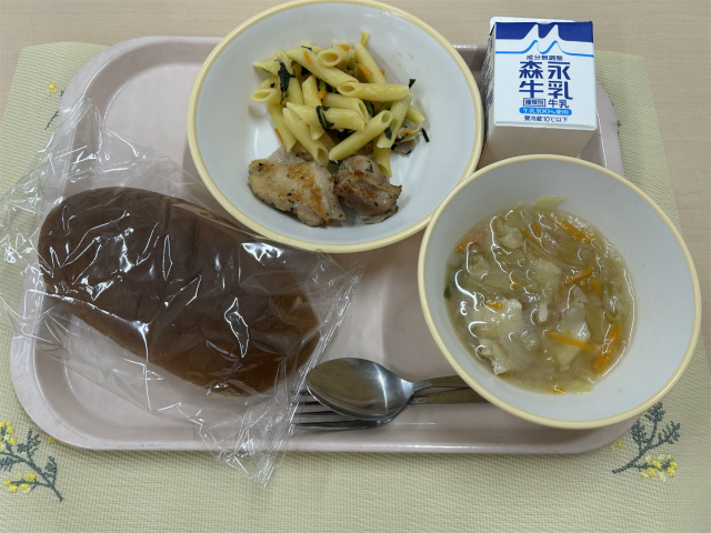 ピンクのトレーに給食が右上から反時計回りで牛乳、小松菜とベーコンのペンネ、鶏肉のハーブ焼き、黒パン、コンソメスープと並べられている写真