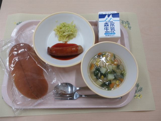 ピンクのトレーに給食が右上から反時計回りで牛乳、ウインナーとせんキャベツ、ソフトフランスパン、野菜のスープ煮と並べられている写真