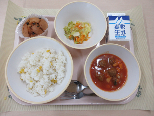 ピンクのトレーに給食が右上から反時計回りで牛乳、春キャベツのソテー、秦野の桜のチョコカップケーキ、コーンピラフ、ミートボールのトマト煮と並べられている写真