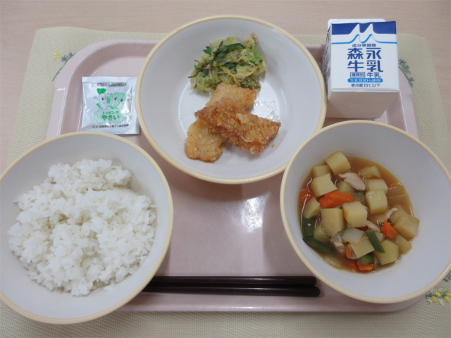 ピンクのトレーに給食が右上から反時計回りで牛乳、いかの吉野揚げとささみとキャベツの昆布和え、やさいふりかけ、ごはん、春野菜の旨煮と並べられている写真