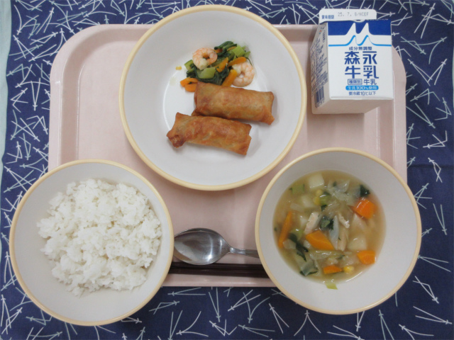 ピンクのトレーに給食が右上から反時計回りで牛乳、春巻きとチンゲンサイの塩炒め、ごはん、野菜のスープ煮と並べられている写真