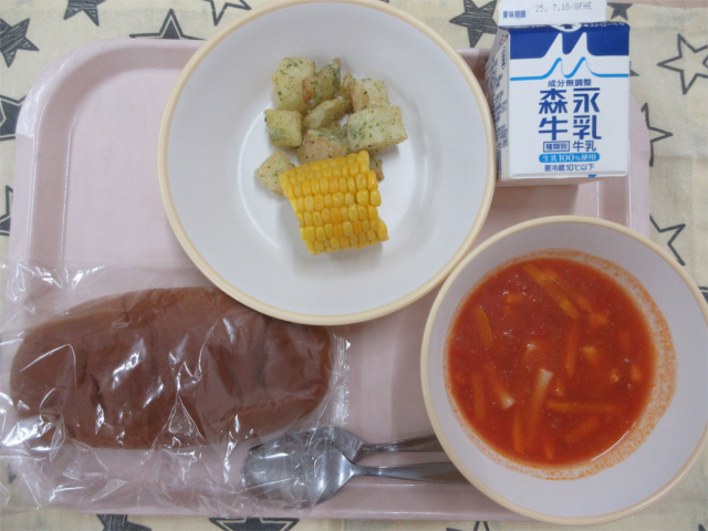 ピンクのトレーに給食が右上から反時計回りで牛乳、青のりポテトととうもろこし、黒パン、ミネストローネと並べられている写真