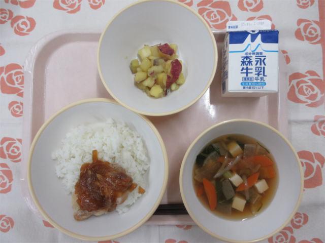 ピンクのトレーに給食が右上から反時計回りで牛乳、さつまいもの甘煮、ノギ屋の鶏飯（ごはん、鶏肉の生姜焼き）、けんちん汁と並べられている写真