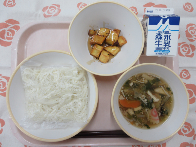 ピンクのトレーに給食が右上から反時計回りで牛乳、大学芋、うどん（ソフト麺）、きのこうどんの具と並べられている写真