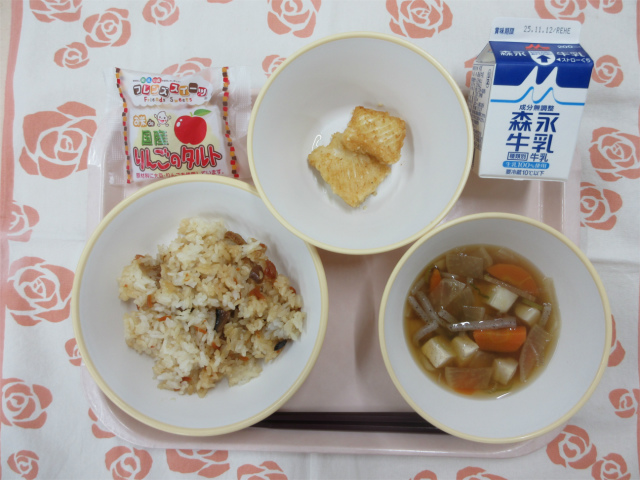 ピンクのトレーに給食が右上から反時計回りで牛乳、いかの吉野揚げ、りんごタルト、かやくごはん、けんちん汁と並べられている写真