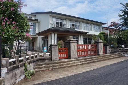 石造りの重厚な門柱と朱色の大きな鉄格子門が特徴的な正面玄関の奥に白い壁の二階建ての校舎の外観写真