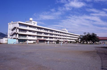 白色で四階建ての大きな学校校舎が建ち、広いグラウンドが広がっている校舎の外観を写した写真