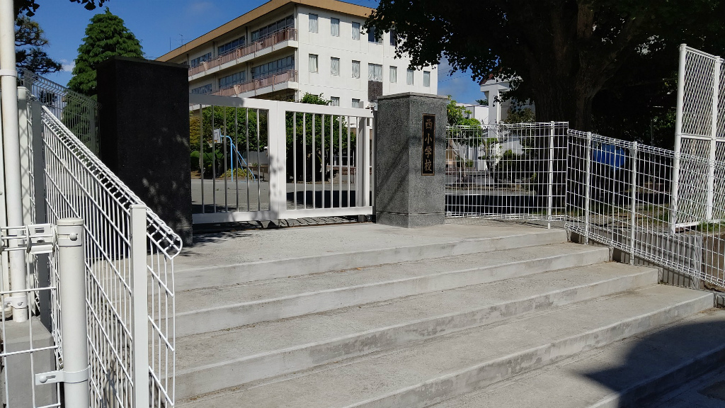 白い鉄柵と門扉で囲まれた学校の正面出入口が写されており、向かって右側の門柱には「西小学校」と刻まれ、奥には校庭と白い校舎が見える写真