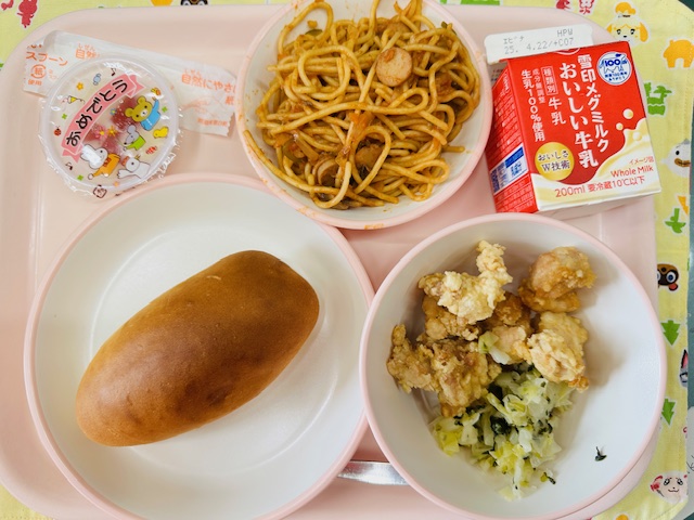 ピンクのトレーに牛乳、スパゲッティナポリタン、デザート、コック帽をかぶった布製マスコット、あじつきパン、鶏肉のからあげとむしやさいと並べられている写真