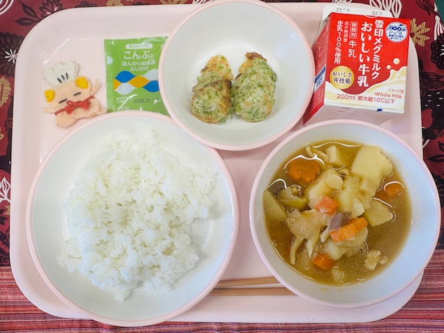 ピンクのトレーに牛乳、ちくわのいそべあげ、抹茶ふりかけ、コック帽をかぶった布製マスコット、ごはん、春野菜のうま煮と並べられている写真