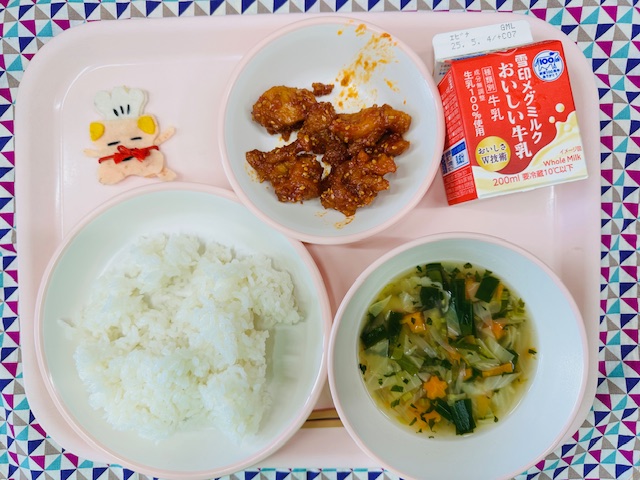 ピンクのトレーに牛乳、ヤンニョムチキン、コック帽をかぶった布製マスコット、ごはん、にらとキャベツのスープと並べられている写真