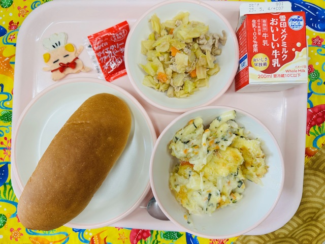 ピンクのトレーに牛乳、はるキャベツのサラダ、いちごジャム、コック帽をかぶった布製マスコット、コッペパン、マロニグラタンと並べられている写真