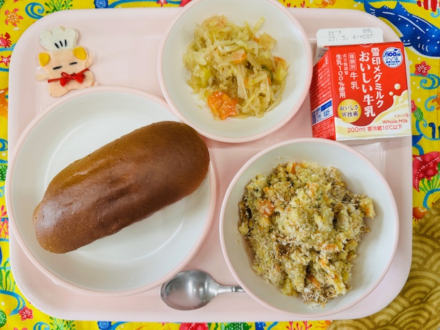 ピンクのトレーに牛乳、ハリハリサラダ、コック帽をかぶった布製マスコット、くろパン、スコップコロッケと並べられている写真