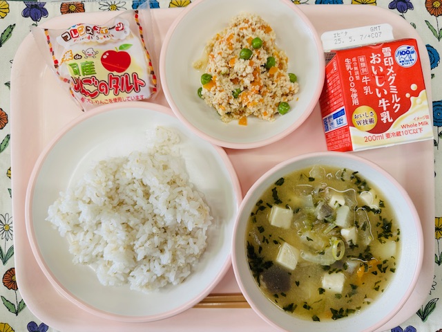 ピンクのトレーに牛乳、とりそぼろ、りんごのタルト、コック帽をかぶった布製マスコット、ごはん、とん汁と並べられている写真