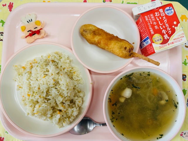 ピンクのトレーに牛乳、アメリカンドッグ、コック帽をかぶった布製マスコット、ベジピラフ、はるさめスープと並べられている写真