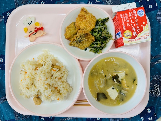 ピンクのトレーに牛乳、アジフライといそかあえ、コック帽をかぶった布製マスコット、たけのこごはん、みそしると並べられている写真