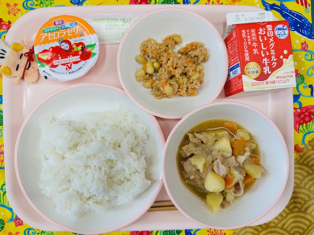 ピンクのトレーに牛乳、ツナだいず、アセロラゼリー、コック帽をかぶった布製マスコット、ごはん、にくじゃがと並べられている写真