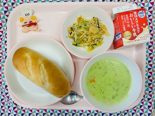 ピンクのトレーに牛乳、ビーフンソテー、コック帽をかぶった布製マスコット、ミルクパン、グリーンスープと並べられている写真