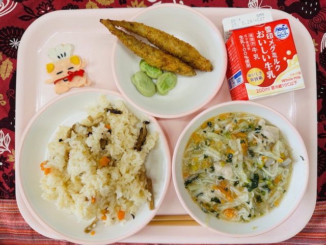 ピンクのトレーに牛乳、ししゃもフライとそらまめ、コック帽をかぶった布製マスコット、ちらしずし、あんかけじると並べられている写真