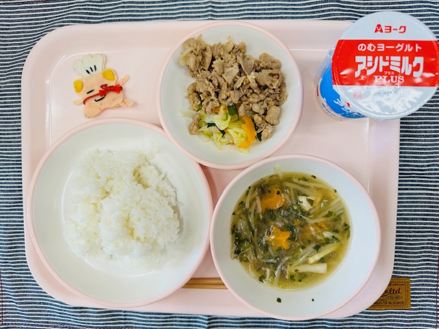 ピンクのトレーにのむヨーグルト、豚肉の生姜焼きとやさいいため、コック帽をかぶった布製マスコット、ごはん、五目はるさめスープと並べられている写真