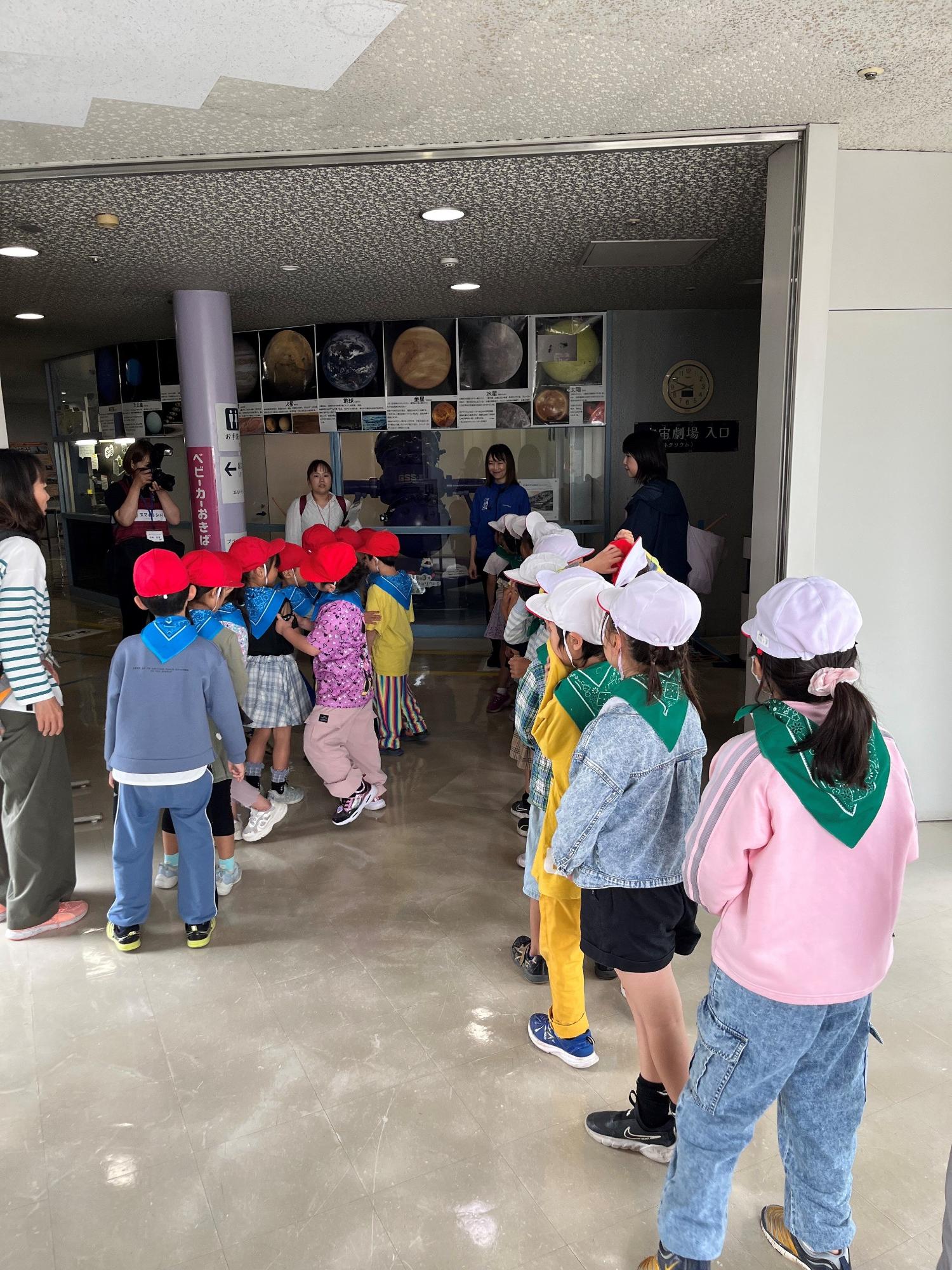 明るいロビーで赤の帽子と青のスカーフを着用したグループと白の帽子と緑のスカーフを着用したグループに分かれた大勢の子どもたちが、引率者とともに列を作り、施設に入場しようとしている様子の写真