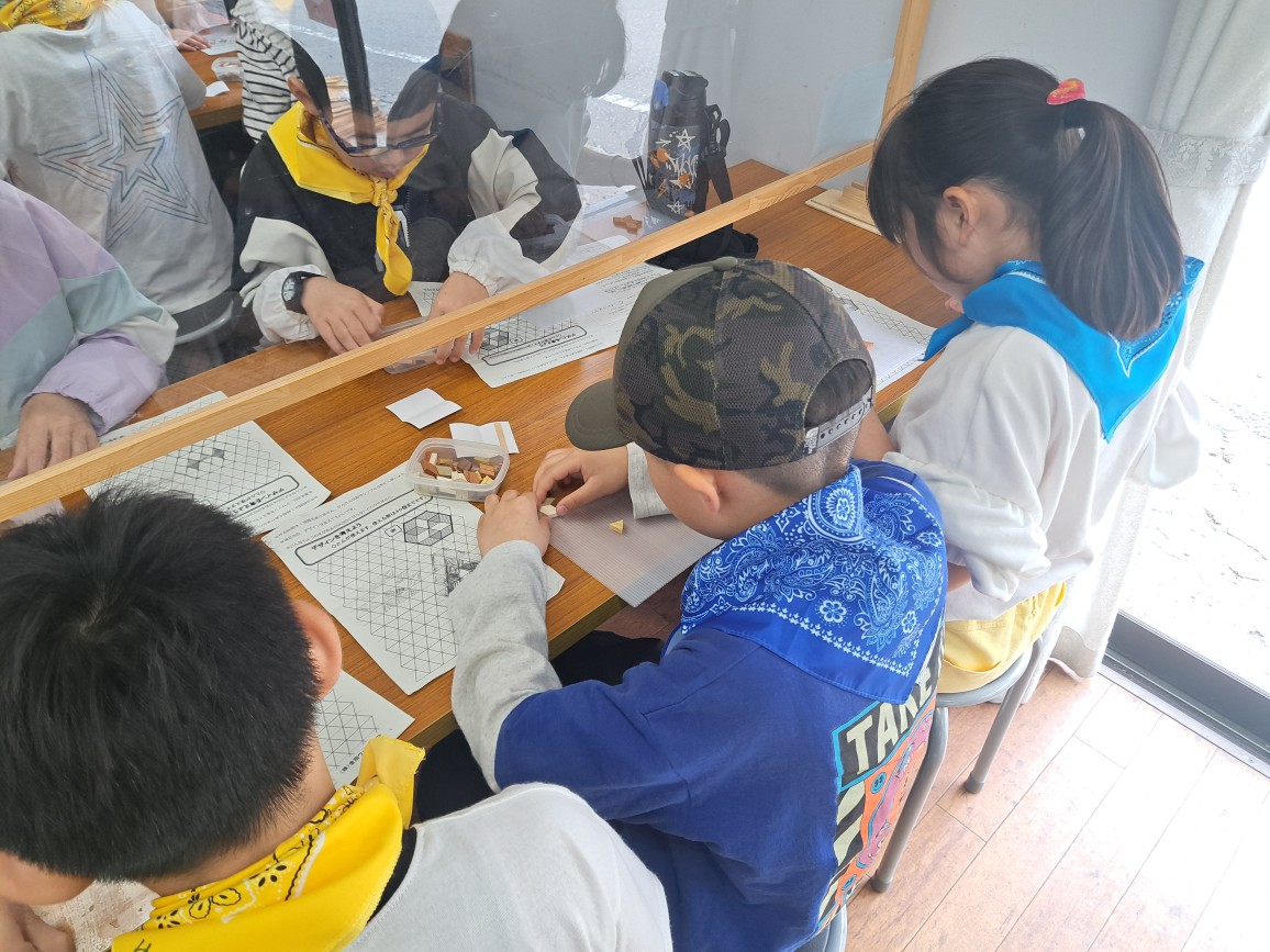 木製の机を囲んでだ数人の子どもたちが、マス目に6角形のコースターのデザインが書かれた図案を見ながら、制作に取り組んでいる様子の写真