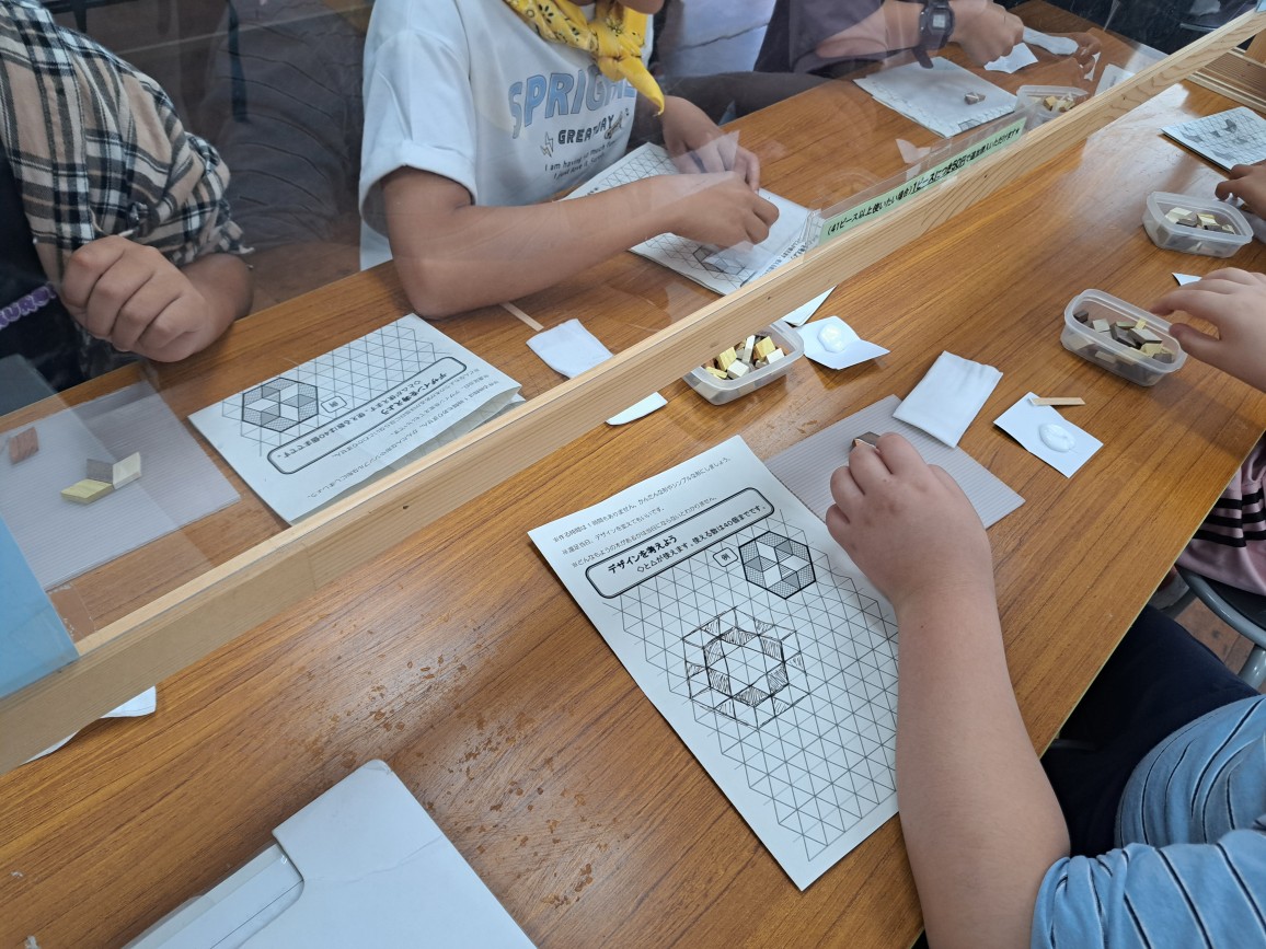 木製の長机の上で、数名の子どもたちが、小さな木片を使い、六角形の模様をデザインする図案を書いたシートを見ながら、コースターの制作に取り組んでいる様子の写真
