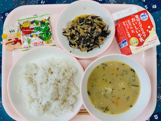 ピンクのトレーに牛乳、ぶたどんの具、入り大豆と昆布、コック帽をかぶった布製マスコット、麦入りごはん、いももちじると並べられている写真