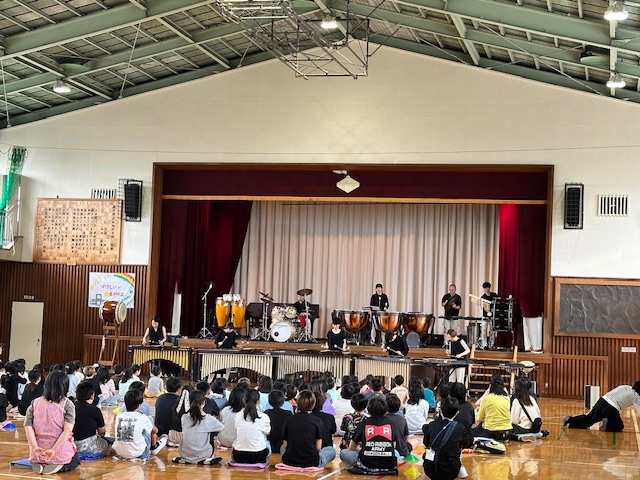 体育館のステージ上にはティンパニやドラムセットなどの打楽器とステージ下には鍵盤打楽器が並べられ、数名の演奏者が演奏を行っており、大勢の子どもたちが座って鑑賞している様子の写真