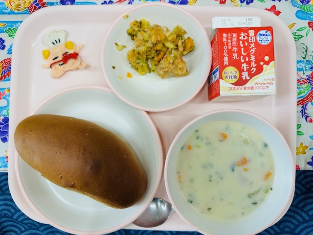 ピンクのトレーに牛乳、とりにくのマリネ、コック帽をかぶった布製マスコット、くろパン、まめシチューと並べられている写真