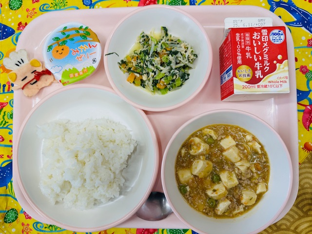 ピンクのトレーに牛乳、とりにくと野菜のナムル、みかんゼリー、コック帽をかぶった布製マスコット、ごはん、マーボーどうふと並べられている写真