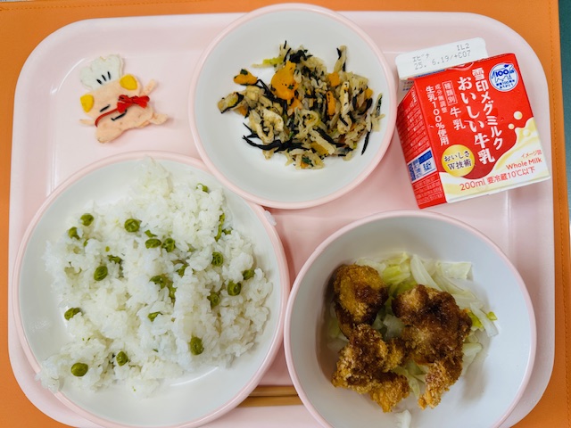 ピンクのトレーに牛乳、切り干し大根の炒め煮、コック帽をかぶった布製マスコット、グリンピースごはん、チキンかつとむしキャベツと並べられている写真