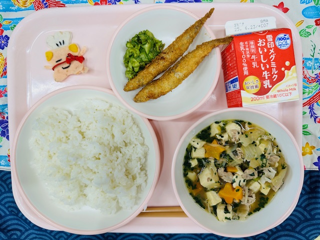 ピンクのトレーに牛乳、ししゃもフライとピリからきゅうり、コック帽をかぶった布製マスコット、ごはん、にくどうふと並べられている写真