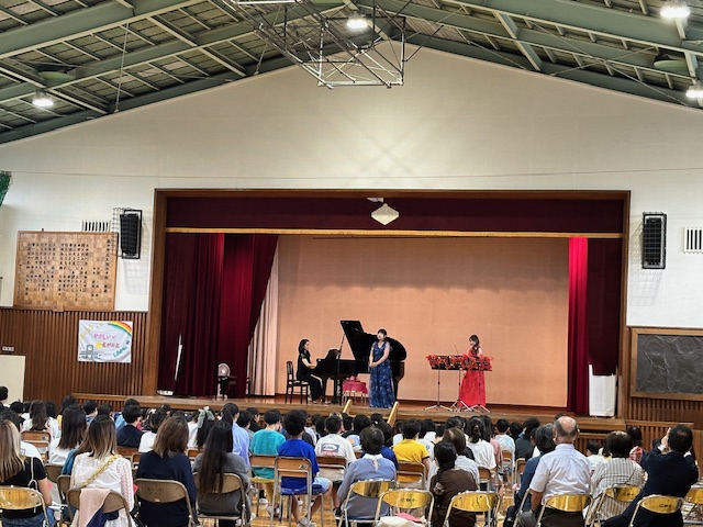 体育館のステージで、黒い服のピアニスト、青いロングドレスのソプラノ歌手、赤いロングドレスのヴァイオリニストの3人の女性が演奏をし、大勢の生徒や関係者が席に座って鑑賞している様子の写真