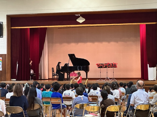 体育館のステージ上での黒い服のピアニストの女性が演奏をし、ステージ下に降りた赤い衣装の女性が観客席に近づきヴァイオリンを演奏している様子の写真