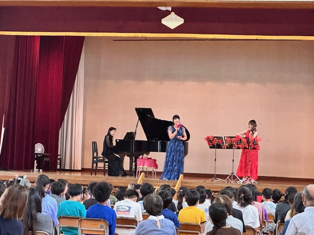 体育館のステージで黒いドレスのピアニスト、赤いドレスのヴァイオリニスト、青いドレスの歌手の女性3人が笑顔で歌い、演奏しており、客席に座った大勢の観客が注目している様子の写真