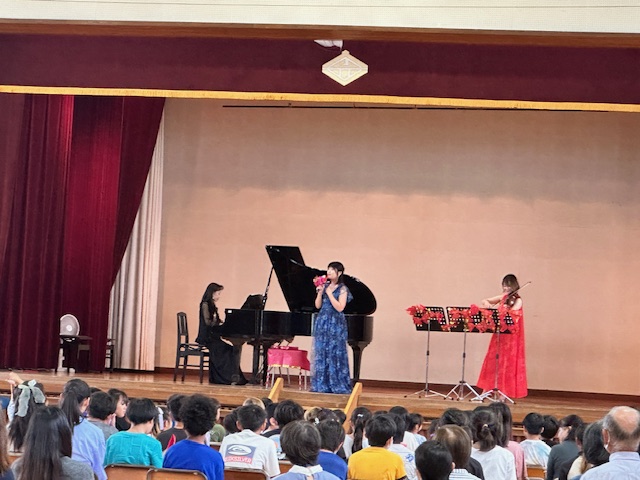 体育館のステージで黒いドレスのピアニスト、赤いドレスのヴァイオリニスト、青いドレスの歌手の女性3人による演奏が行われ、客席に座った大勢の観客が注目している様子の写真