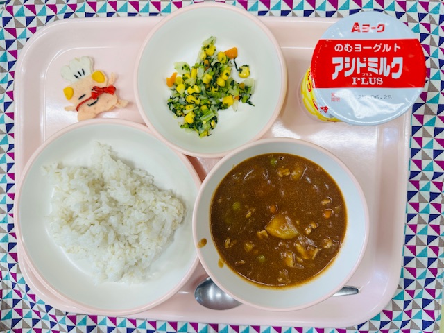 ピンクのトレーにでアシドミルク、野菜ソテー、コック帽をかぶった布製マスコット、ごはん、ハヤシライスと並べられている写真