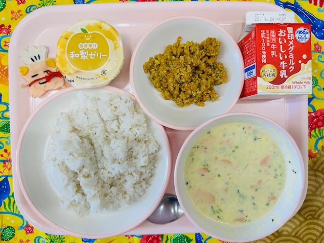 ピンクのトレーに牛乳、ドライカレー、和梨ゼリー、コック帽をかぶった布製マスコット、ごはん、コーンスープと並べられている写真