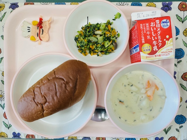 ピンクのトレーに牛乳、あおないため、コック帽をかぶった布製マスコット、くろパン、しろはなまめいりシチューと並べられている写真