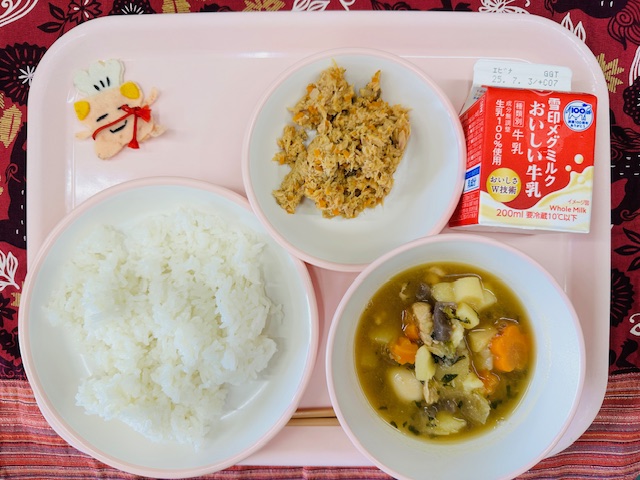 ピンクのトレーに牛乳、ツナそぼろ、コック帽をかぶった布製マスコット、ごはん、やさいのうま煮と並べられている写真