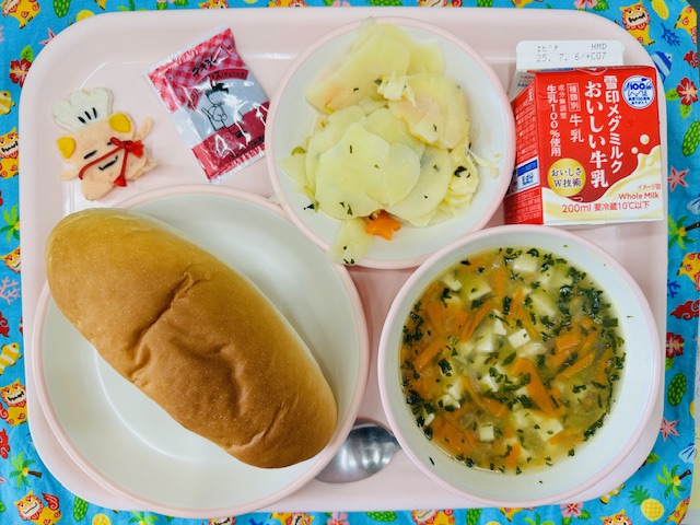 ピンクのトレーに牛乳、じゃがいものチーズ焼き、チョコだいず、コック帽をかぶった布製マスコット、コッペパン、コーン中華スープと並べられている写真