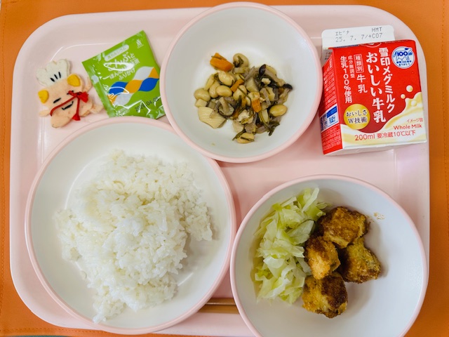 ピンクのトレーに牛乳、ごもくまめ、ふりかけ、コック帽をかぶった布製マスコット、ごはん、白身魚ふらいとむしキャベツと並べられている写真