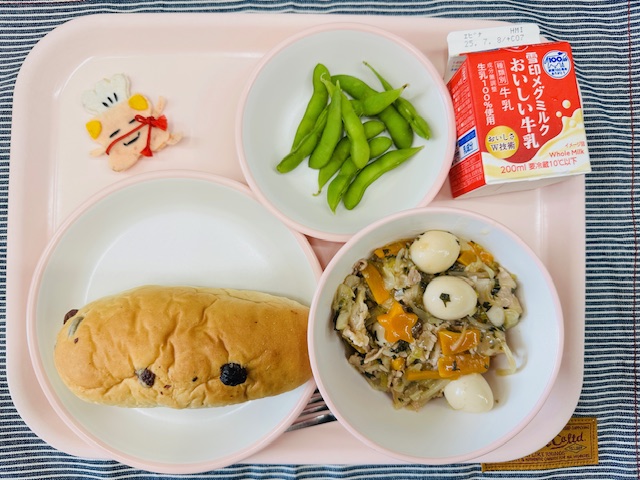 ピンクのトレーに牛乳、えだまめ、コック帽をかぶった布製マスコット、ぶどうパン、やきそばあんかけと並べられている写真